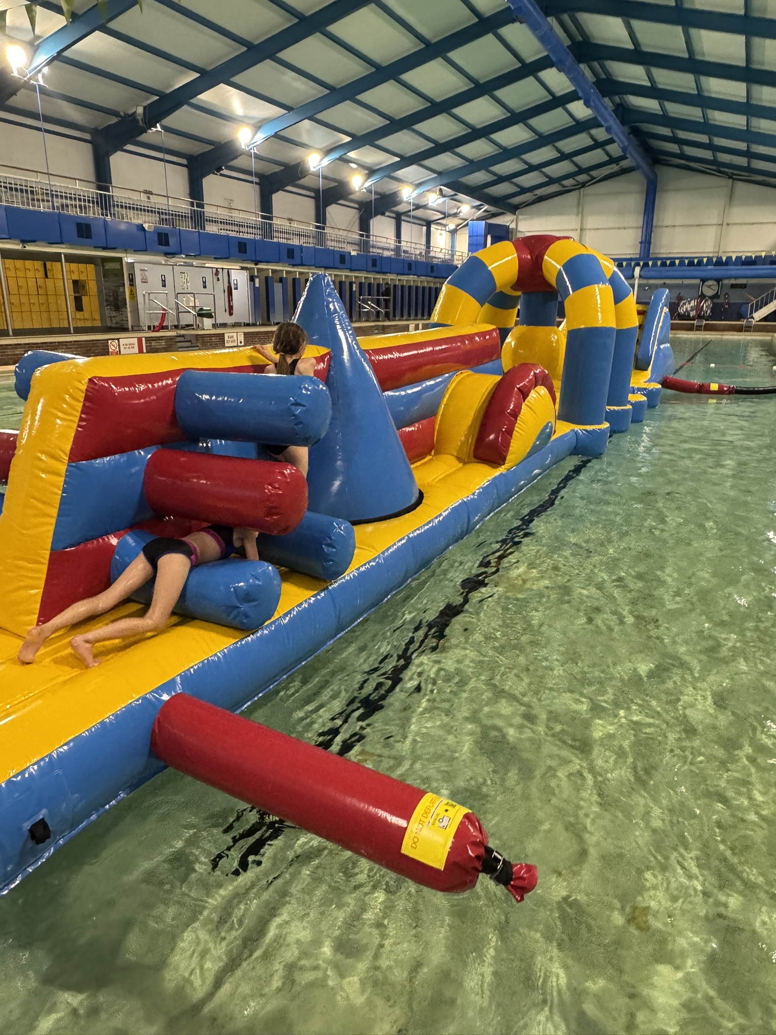 Child navigating a colorful inflatable obstacle course in an indoor swimming pool. The vibrant yellow, blue, and red structure evokes fun and playfulness.