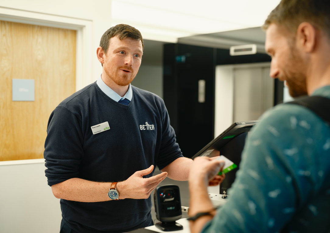 image shows a man in a dark blue jumper wearing a name badge assisting another person with information.