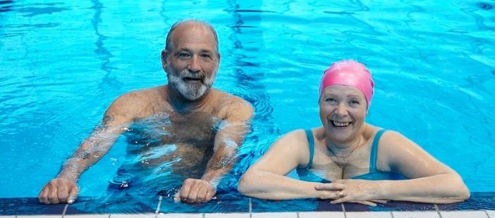 image shows a senior male and female smiling in the water at the edge of a swimming pool.