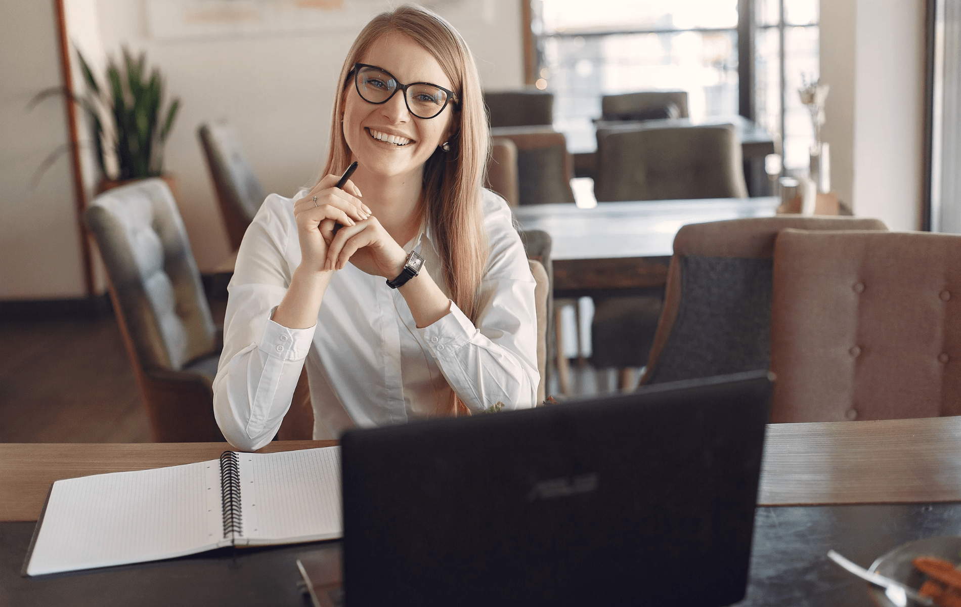 A freelancer smiling at a table with her work laptop, she has a watch on to show the flexibility of her time and schedule. 