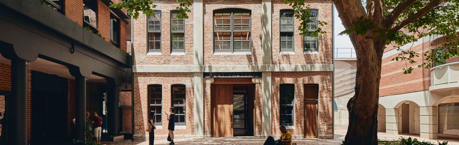 A multi-story red brick building with large windows and a wooden door, partially shaded by a large leafy tree.