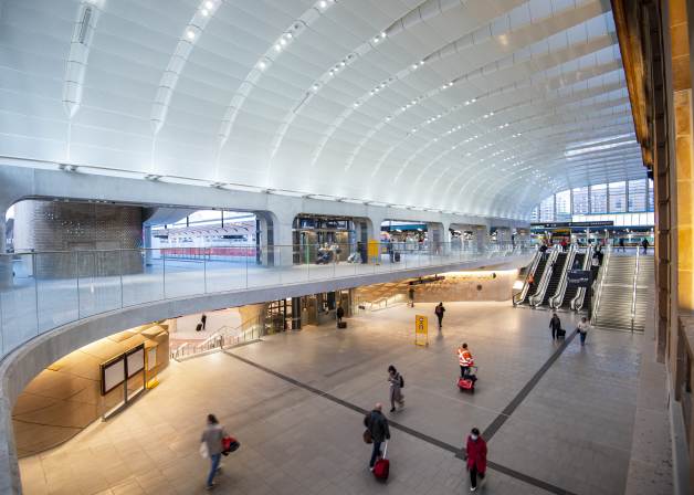 Station concourse interior with a white, high arched ceiling, four escalators, a staircase, and people walking with luggage on multiple levels. At the back of the image is a metro train.