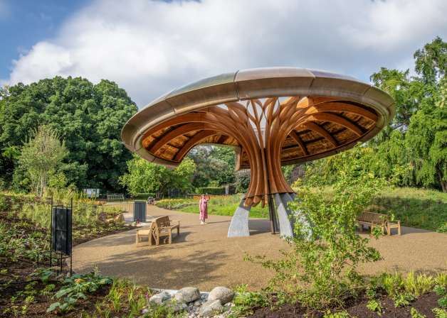 A large, mushroom-shaped wooden and metal structure stands in a garden with trees and a person walking.