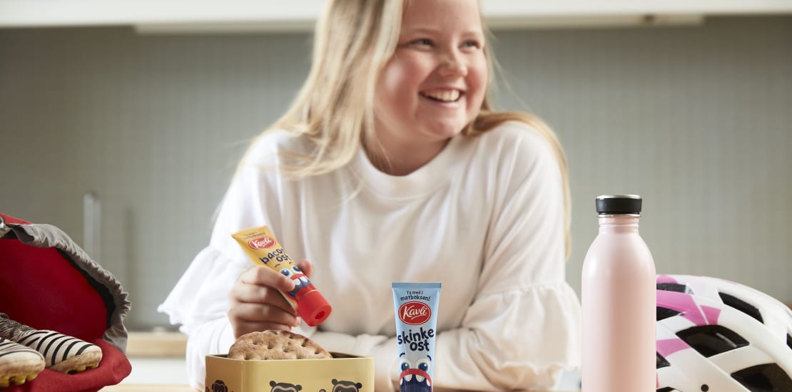En smilende ung jente pakker matpakken sin til skoledagen. Hun holder en tube med Kavli BaconOst, som får plass i matboksen.