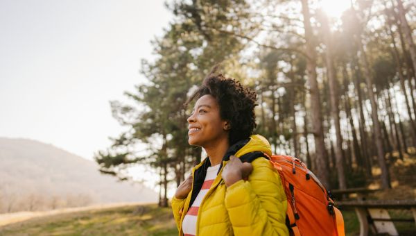Young Black woman with yellow jacket and orange backpack, smiling and hiking outdoors.