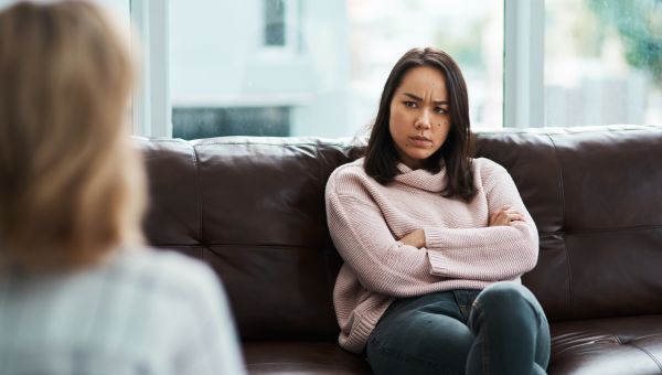 a young woman sits on a couch during a therapy session, arms folded, with an angry look on her face