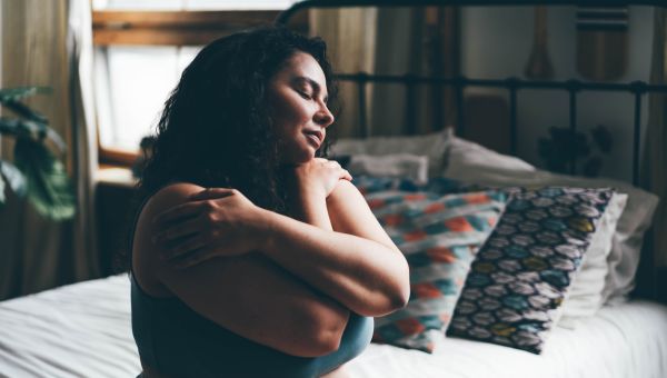 a woman wearing underwear sits on the edge of her bed with her eyes closed, enfolding herself in a self-embrace