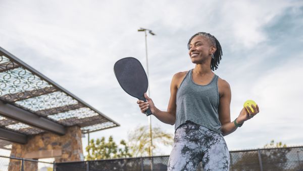 a fit middle aged Black woman in workout wear smiles as she holds a pickleball racket and ball
