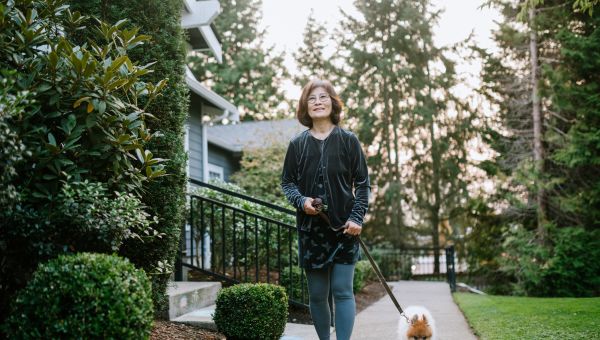 an older Asian American woman walks a dog down a suburban sidewalk 