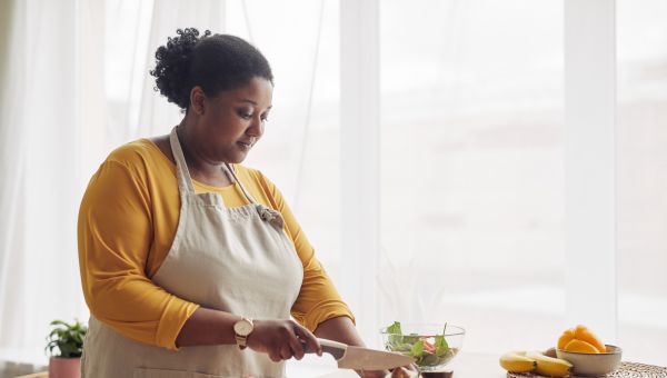 an overweight Black woman slices an avocado while she makes a healthy meal in a sunny kitchen