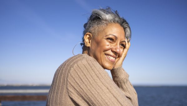 a middle aged Black woman smiles and looks off to sea on a sunny day near the beach