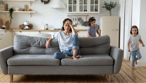 stressed mom sits on couch at home while young daughter and son run around the living room