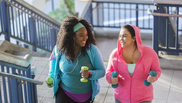 two overweight young women, a Black woman and a white woman, chat amiably as they exercise by walking up stairs holding light dumbbells