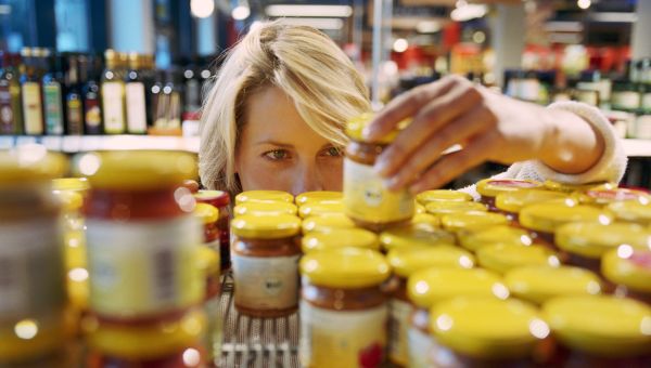 a white woman with blonde hair peers at a shelf of jarred condiments, reading the nutrition labels to find out how much added sugar they contain