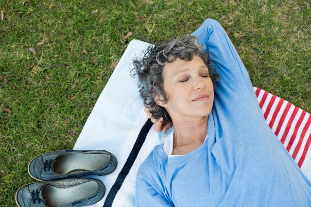 Smiling senior woman lying on mat at garden with eyes closed. High angle view of beautiful mature woman relaxing after yoga in park. Top view of senior woman sleeping at park.