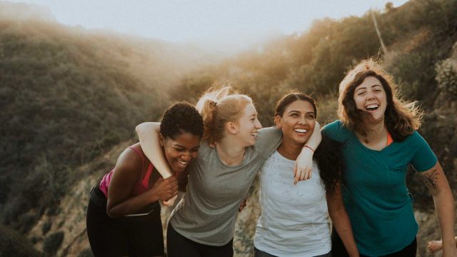 Four women friends are building healthy relationships as they laugh while hiking arm in arm.