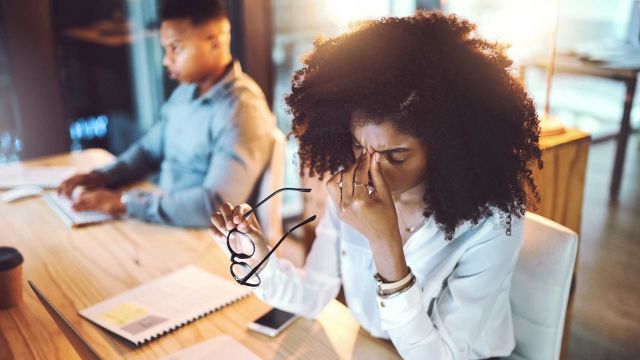 Stressed out woman at work holds her glasses in one hand and pinches the bridge of her nose with her other.