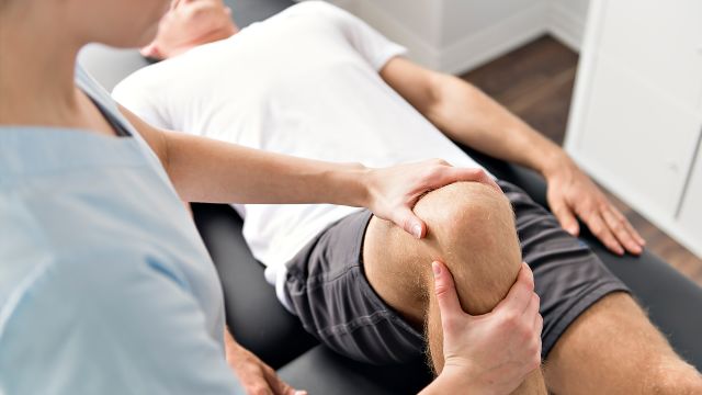 A man with knee pain lies in a table while a physical therapist helps with knee-friendly leg exercises.