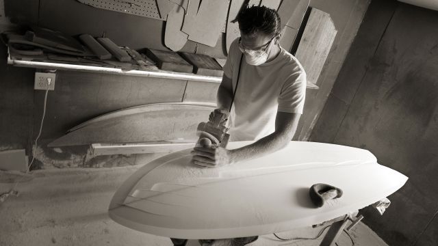 Man shapes a surfboard in a work area.