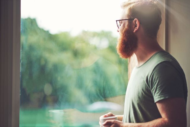 Young adult with a full red beard looks pensively out a window
