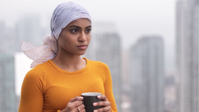 A young woman with breast cancer wears a scarf over her head. She is holding a mug of coffee.