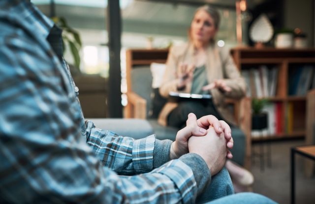 a male patient sits in a therapists office; we see the man's hands in the foreground and his female therapist in the background