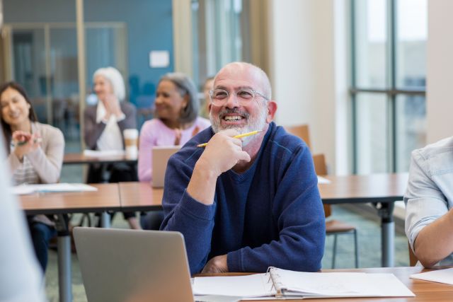 a smiling white middle aged man with a beard sits in an adult learning class as he enjoys the lesson