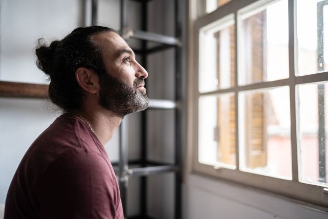 a middle-aged bearded white man with a ponytail gazes out a window with a smile