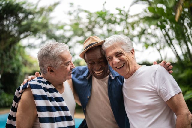 A group of three older adult male friends, two Latino men and one Black man, stand close together sharing a laugh.