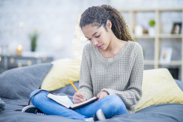 teen girl with adhd writes into a journal about her negative thoughts while sitting cross legged on her bed.