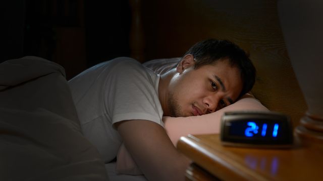 A young man with anxiety struggles with insomnia and looks at the clock while trying to sleep.