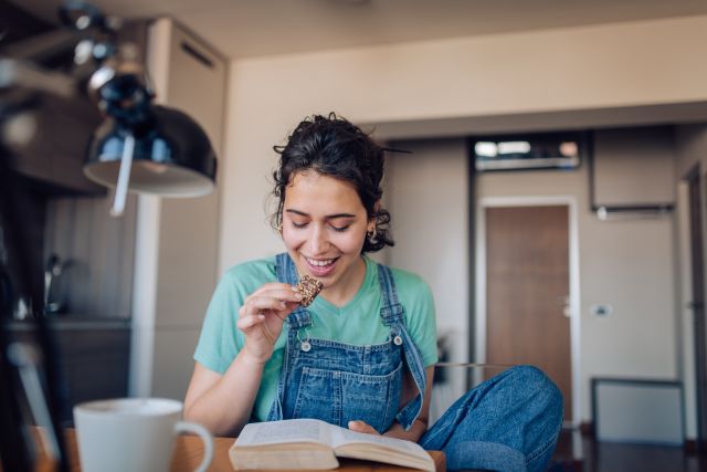 woman sitting at her desk reading a book, eating a chocolate covered cookie