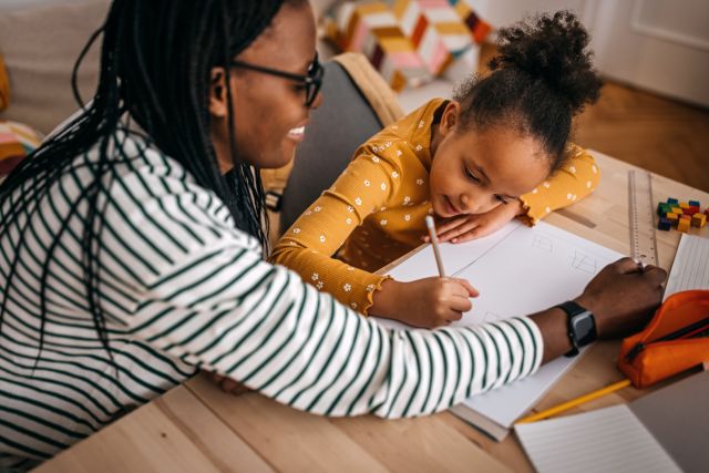 smiling mother assisting daughter in writing homework on table at home