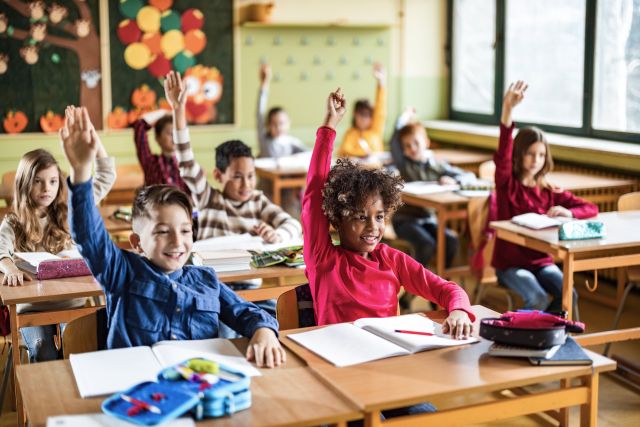 a diverse elementary school classroom with Black and white girls in the front row of desks raising their hands to answer a question 