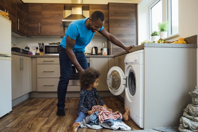 A Black man and a Black child gather clothing to put into a washing machine.