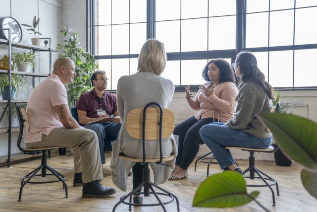 Multiethnic group of adults sit on chairs in a circle to discuss their mental health, addictions, and struggles in a group setting with a professional leading the discussion.