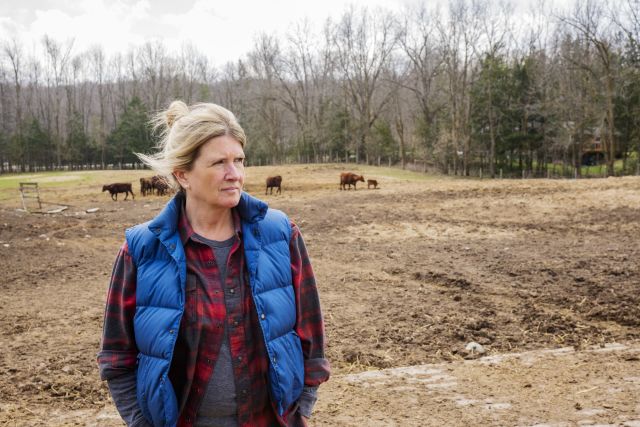 a middle aged white woman in a rural area stands in a cow field