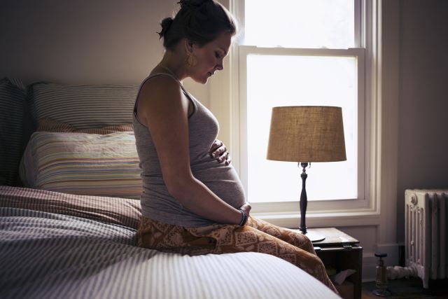 a young white pregnant women sits in a dimly lit room near a window, placing her hands on her belly