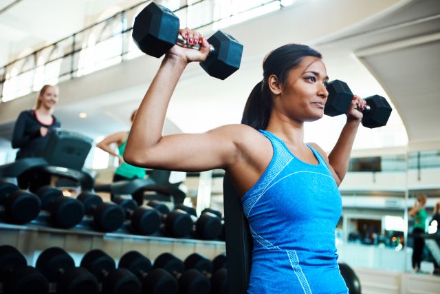 A young woman sits upright in a gym and does a set of seated shoulder presses with dumbbells.