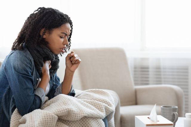 Black adult woman sitting on a sofa under a blanket coughing