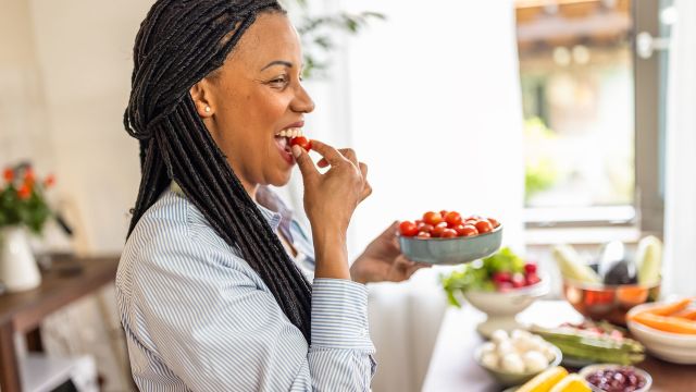 close up image of a woman smiling and eating a cherry tomato while holding a bowl of tomatoes
