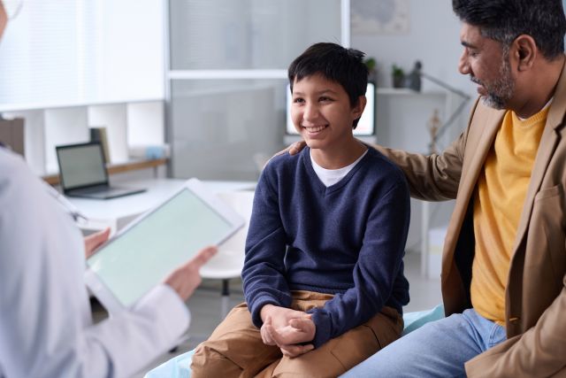 a dark-haired preteen boy sits and smiles in the doctor's office as he and his father discuss HPV vaccination with their healthcare provider