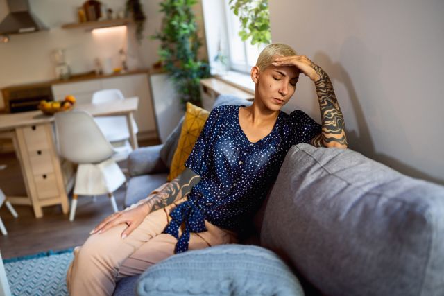 a middle aged white woman with close-cropped blonde hair and tattoos along her arms sits on a couch with her left hand on her head, feeling heavy fatigue that may be an early sign of a hart attack