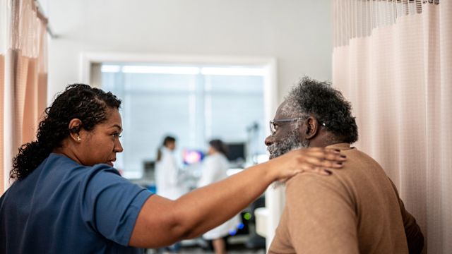 A healthcare provider leads a senior male patient into the exam room at the start of an appointment to follow up on hep C treatment.