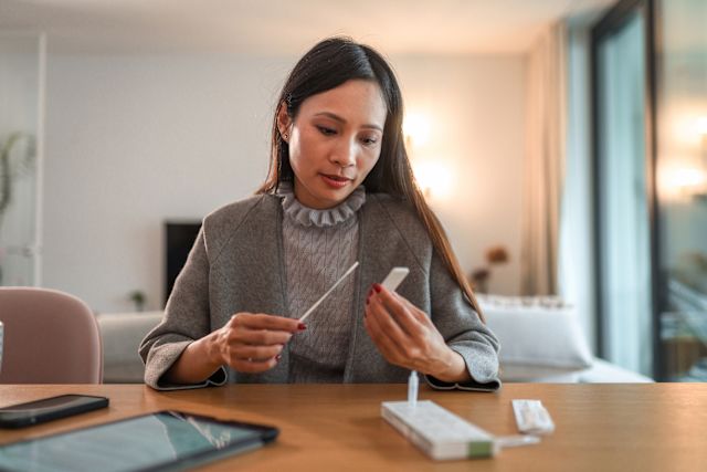 a middle aged Asian woman sits at a table in her home with a COVID antigen test, preparing to test to find out whether she has COVID