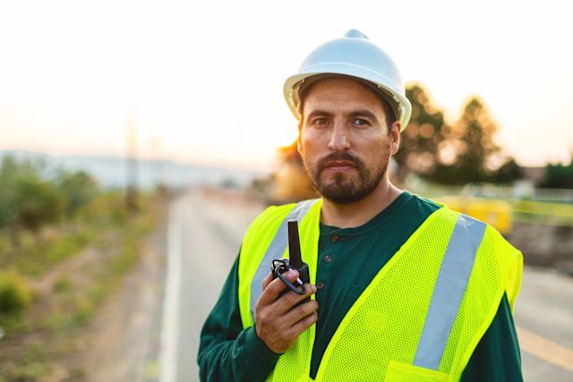 A middle-aged Latino man wears a yellow vest while doing outdoor work on a highway. He has a worried look on his face because he is concerned about the air quality resulting from air pollution. 