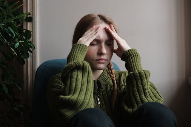 a young white woman with red hair wearing a green sweater sits on the floor with her hands on her head as she experiences a headache caused by COVID