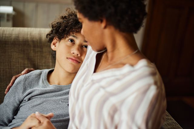 Adult woman talking with male school aged child on sofa, offering comfort with arm around his shoulder.