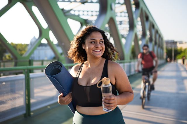 adult woman of color walking outside in workout clothes carrying a yoga mat
