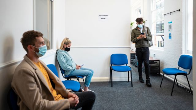 Several adult patients with primary immunodeficiency wear masks as they sit or stand in a waiting room at a healthcare provider's office.